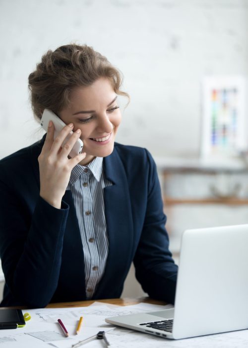 Portrait of beautiful happy smiling young architect woman talking on phone and working on laptop at home office desk. Attractive cheerful model using smartphone and computer, making call, taking order